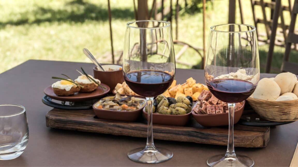 Two glasses of red wine are in front of a board of cheese, olives, and other wine-tasting nibbles.