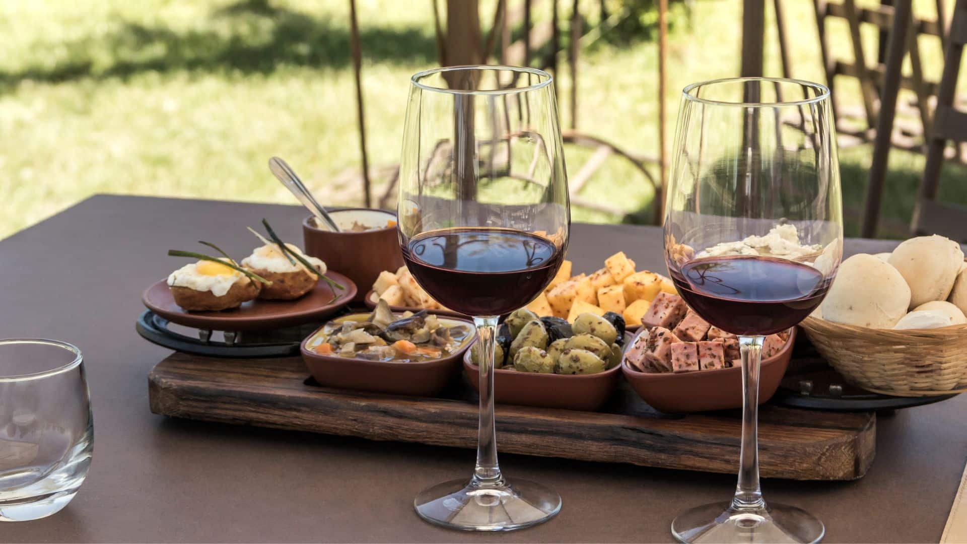 Two glasses of red wine are in front of a board of cheese, olives, and other wine-tasting nibbles.