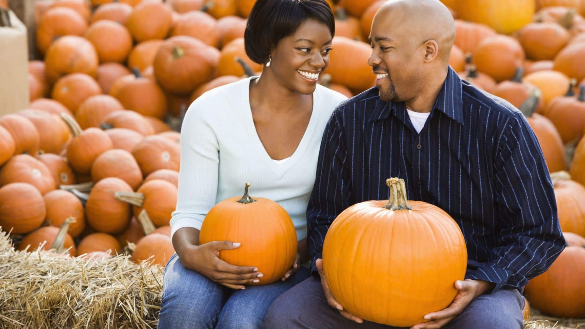 a couple sits on a hay bale with pumpkins in their laps and pumpkins behind them