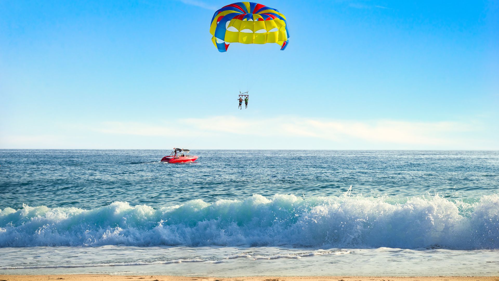 Two people are in the air on a parasail, pulled by a red boat close to the ocean shore.